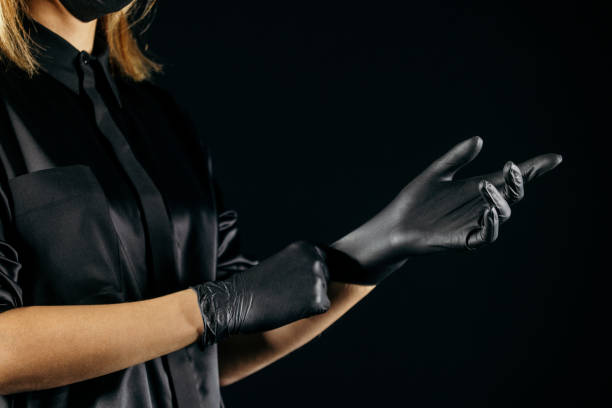 Woman is preparing for her job and putting on black protective gloves. She is wearing black uniform. Photo taken with dark background. She is professional permanent make up artist